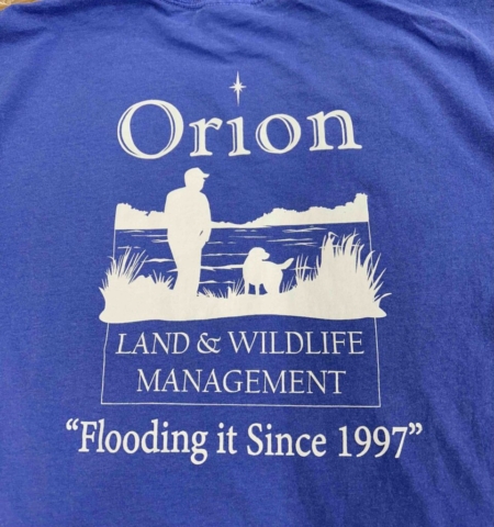 Orion Land and Wildlife Management Cobalt blue shirt with white screen print of person and dog silhouette by water's edge. Printed text reads Orion Land and Wildlife Management - Flooding it since 1997.